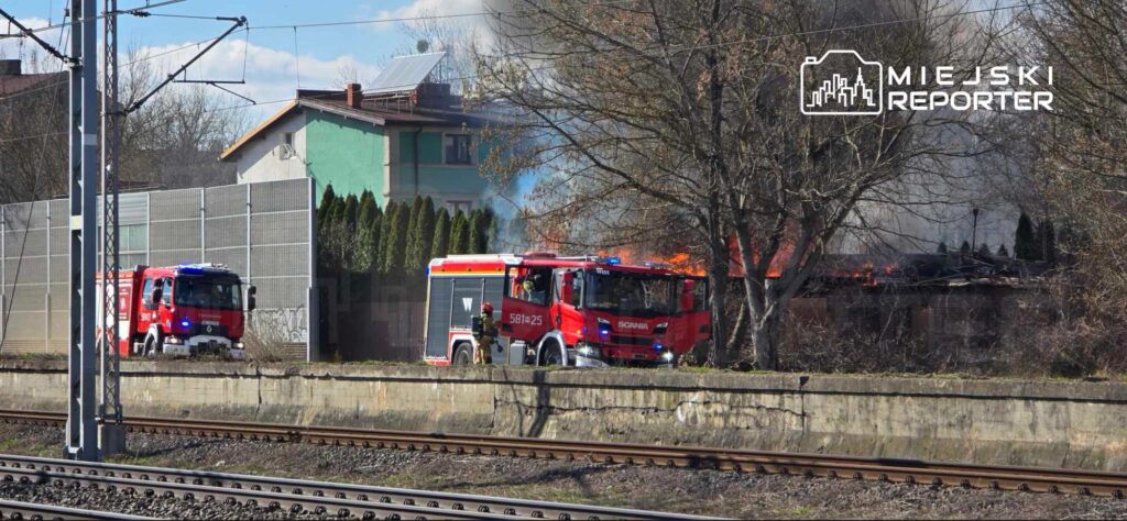 Czerwony wóz strażacki z włączonymi sygnałami podjeżdża do płonącego budynku w pobliżu torów kolejowych.