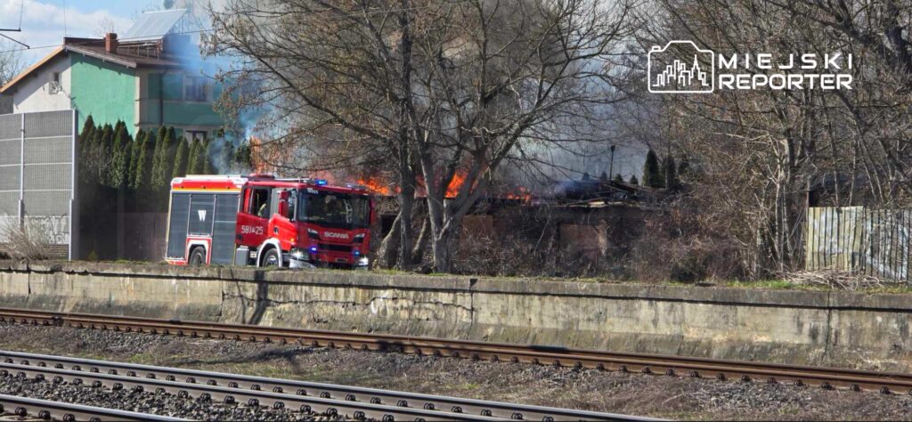 Straż pożarna z włączonymi sygnałami dojeżdża do pożaru w pobliżu torów kolejowych, w tle widoczne płomienie.
