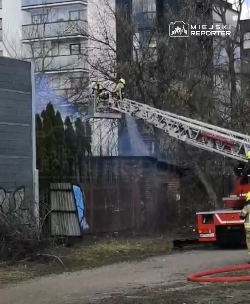 Strażacy na drabinie gaśniczej polewają wodą teren w pobliżu budynku mieszkalnego.