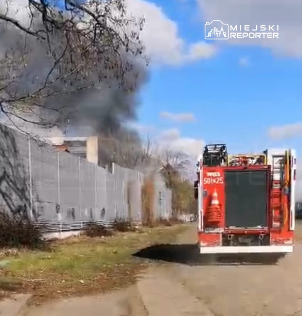 W tle widoczny dym unoszący się z budynku, a na pierwszym planie widać wóz strażacki na drodze.