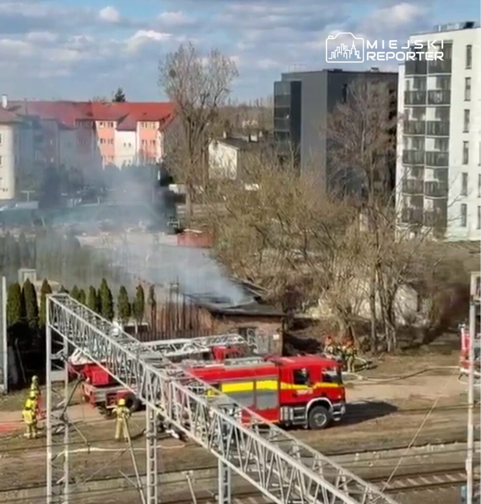 Strażacy gaszą pożar w budynku, obok stoi wóz strażacki, w tle widać mieszkalne bloki i dym unoszący się z płonącego obiektu.