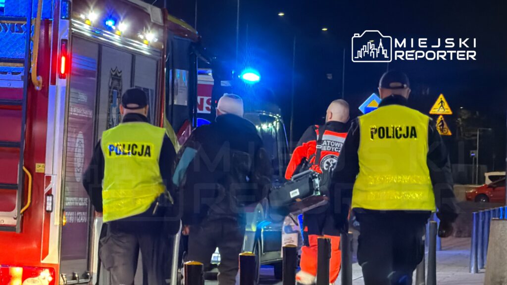 Grupa ratowników medycznych i policjantów w odblaskowych kamizelkach kieruje się w stronę karetki na oświetlonej ulicy.