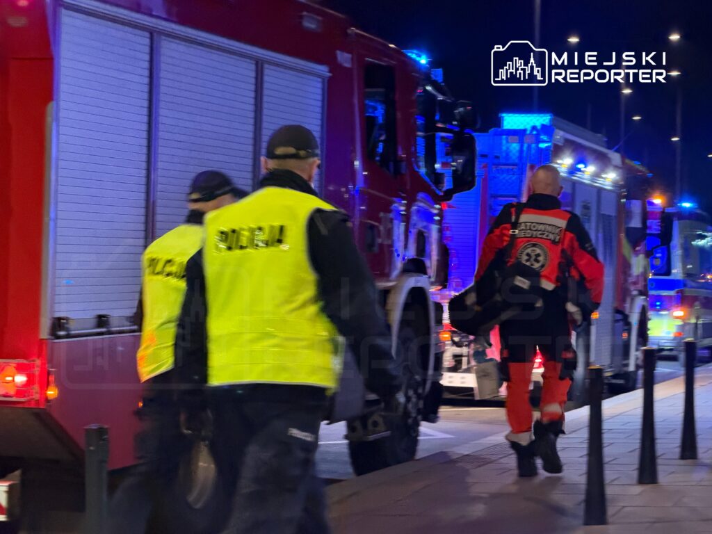 Grupa ratowników i policjantów w odblaskowych kamizelkach przemieszcza się w kierunku wozów strażackich w nocy.