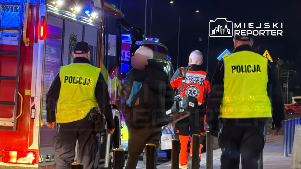 Grupa służb ratunkowych, w tym policjanci w kamizelkach odblaskowych oraz ratownik medyczny, pracuje przy wozie strażackim w nocy.