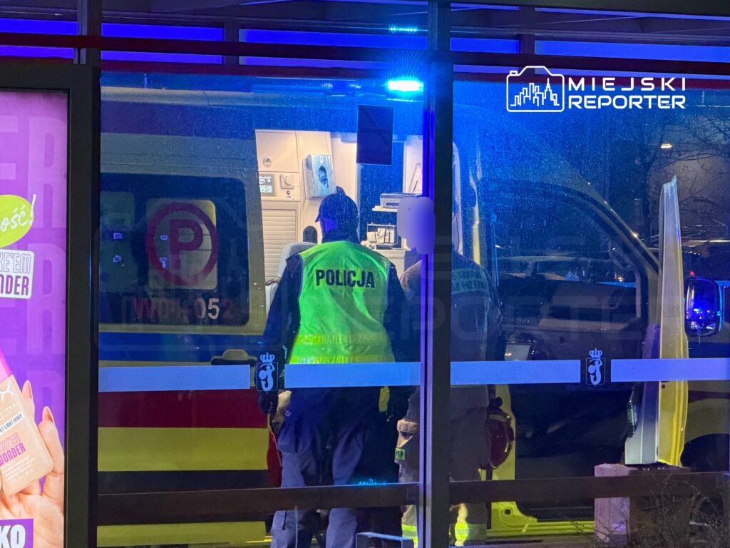 Policjanci w kamizelkach odblaskowych rozmawiają z pracownikami karetki w oświetlonym miejscu.