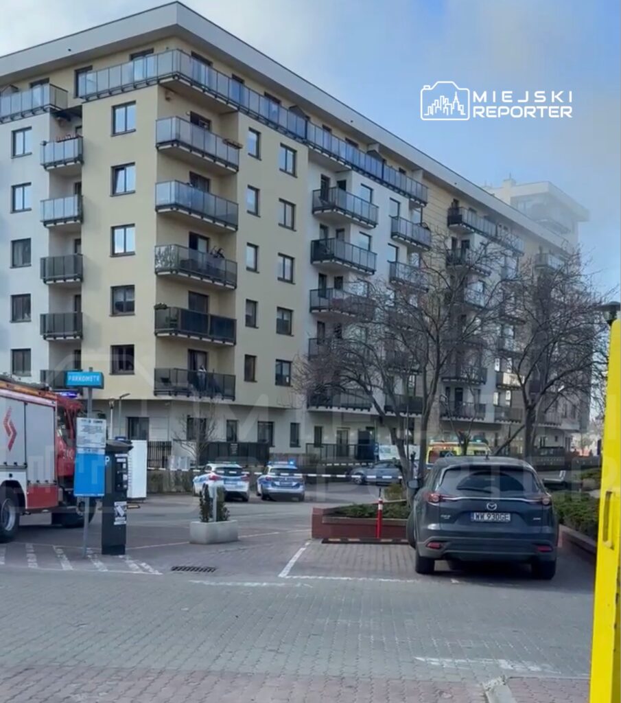 Straż pożarna oraz radiowozy policyjne zaparkowane przed nowoczesnym budynkiem mieszkalnym w miejskiej okolicy.