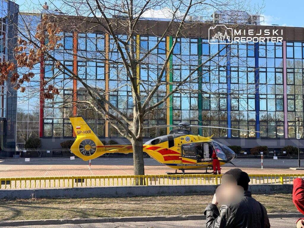 Żółty helikopter z logo HEMS ląduje na parkingu przed nowoczesnym budynkiem z kolorowymi szybami.
