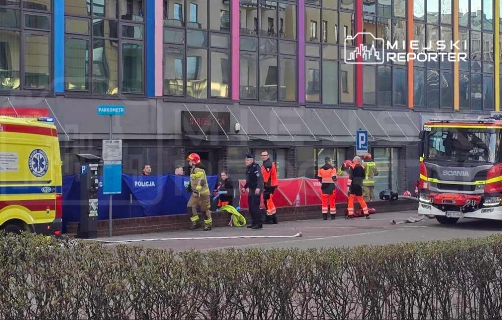 Grupa ratowników oraz policjantów pracuje na miejscu zdarzenia przed nowoczesnym budynkiem, obok karetki i wozu strażackiego.