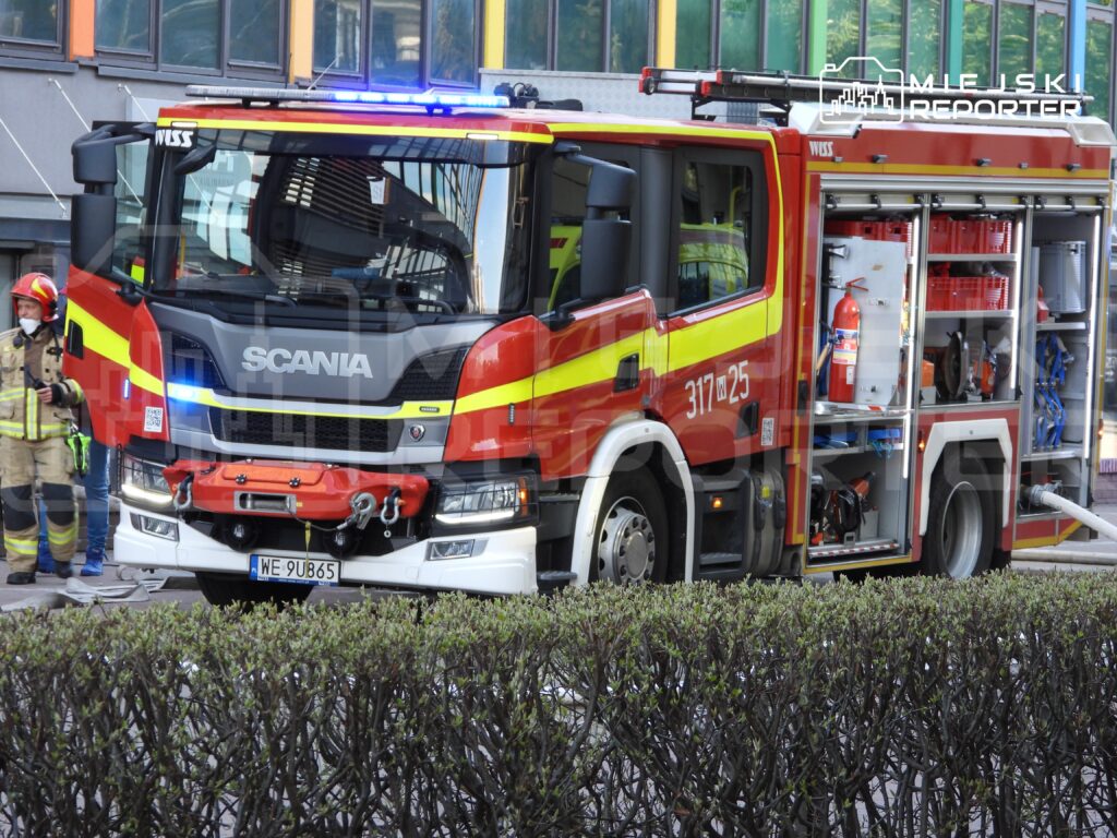 Wóz strażacki marki Scania z włączonymi sygnalizacjami świetlnymi stoi na ulicy obok strażaka w mundurze.