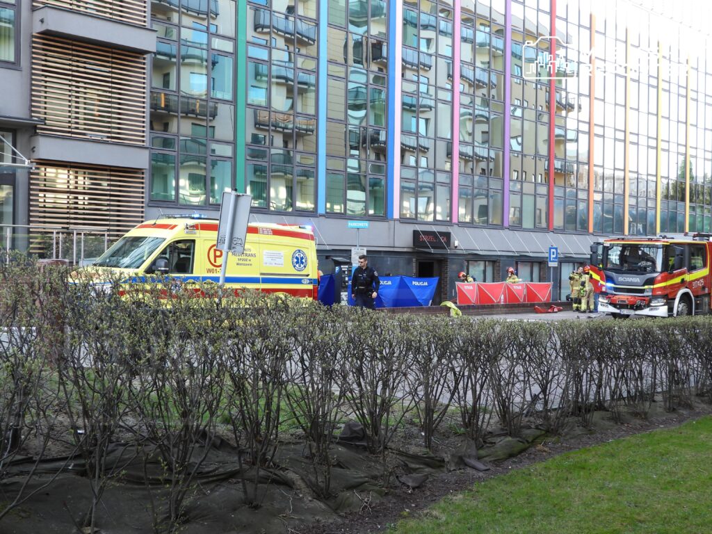 Ambulans i wóz strażacki zaparkowane przed nowoczesnym budynkiem, w tle policjanci zabezpieczający teren.