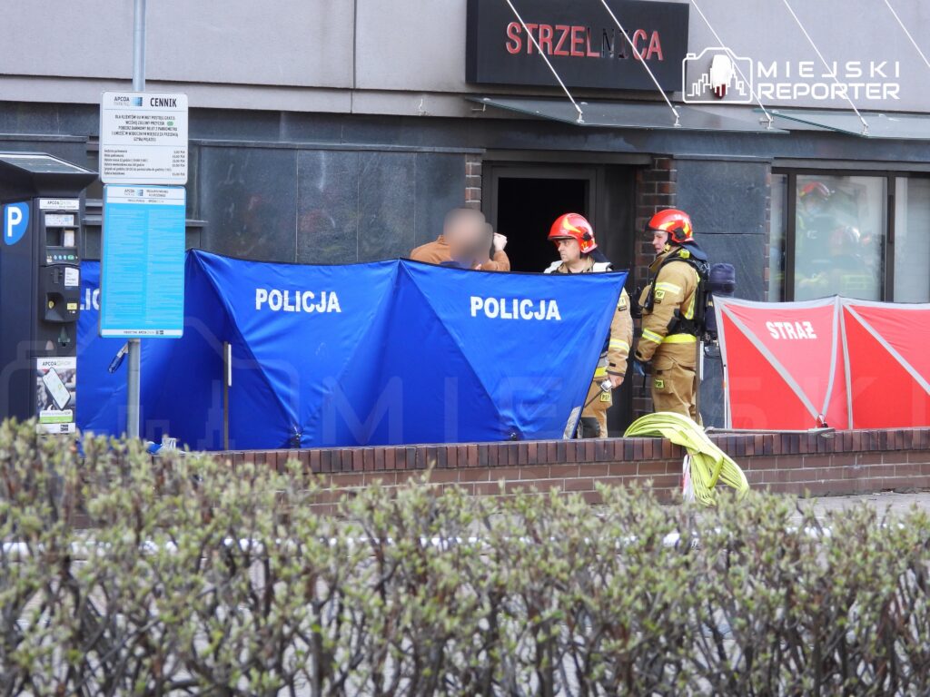 Grupa strażaków i policjantów rozmawia przed budynkiem, przy którym ustawione są namioty zabezpieczające.