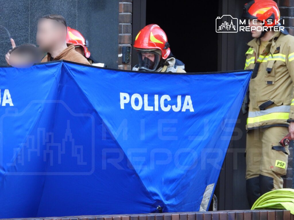 Mężczyzna w brązowej kurtce stoi obok niebieskiego namiotu policyjnego, a w tle widać strażaków w uniformach.