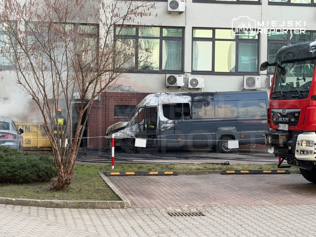 Strażak w hełmie gasi pożar w pobliżu spalonego vana przy nowoczesnym biurowcu, z widocznymi smugami dymu.