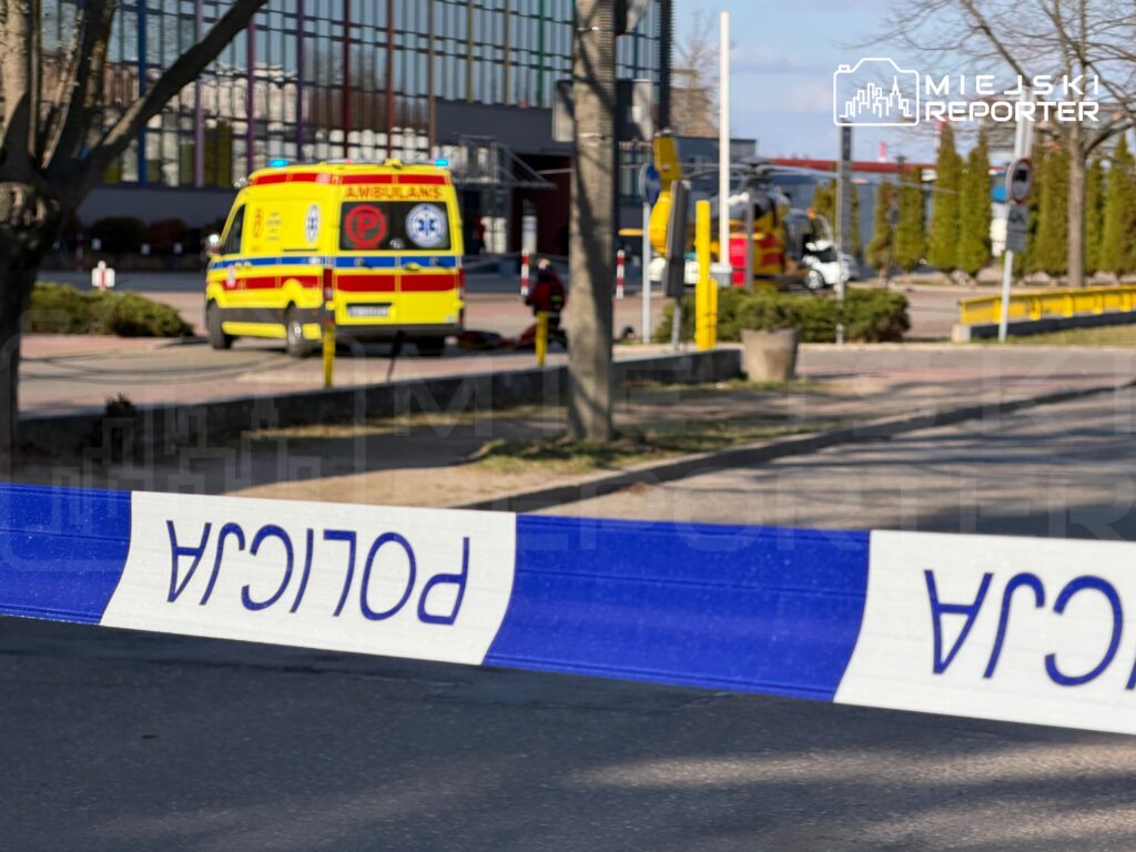 Karetka pogotowia stojąca za policyjną taśmą zabezpieczającą na terenie miejskim.