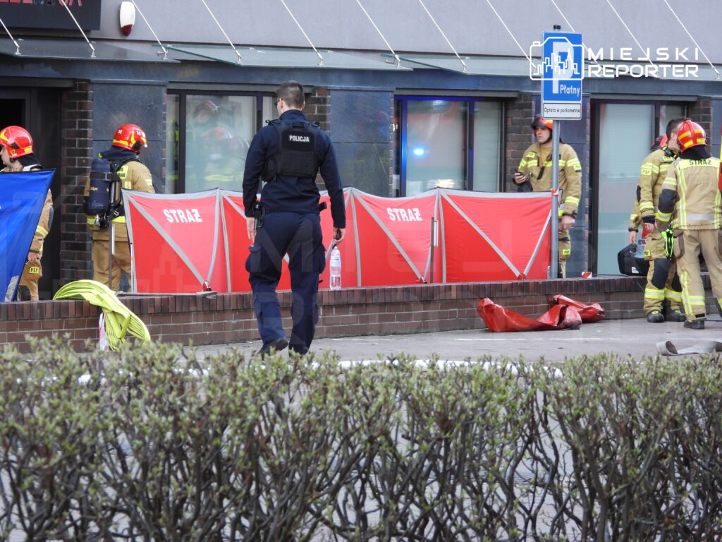 Grupa strażaków w hełmach i mundurach pracuje przy budynku, obok nich policjant w mundurze obserwuje sytuację.