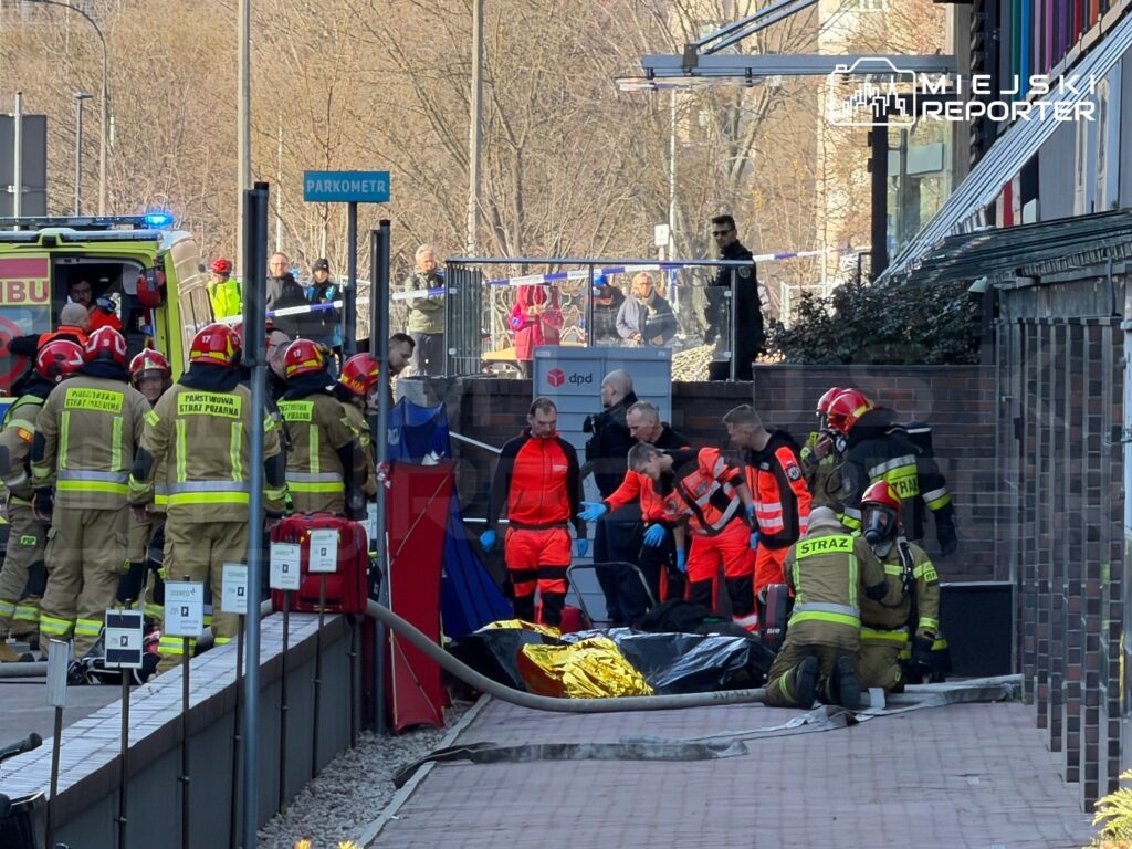 Ratownicy medyczni i strażacy udzielają pomocy poszkodowanemu na chodniku przed budynkiem, w otoczeniu pojazdów ratunkowych.
