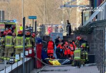 Ratownicy medyczni i strażacy udzielają pomocy poszkodowanemu na chodniku przed budynkiem, w otoczeniu pojazdów ratunkowych.