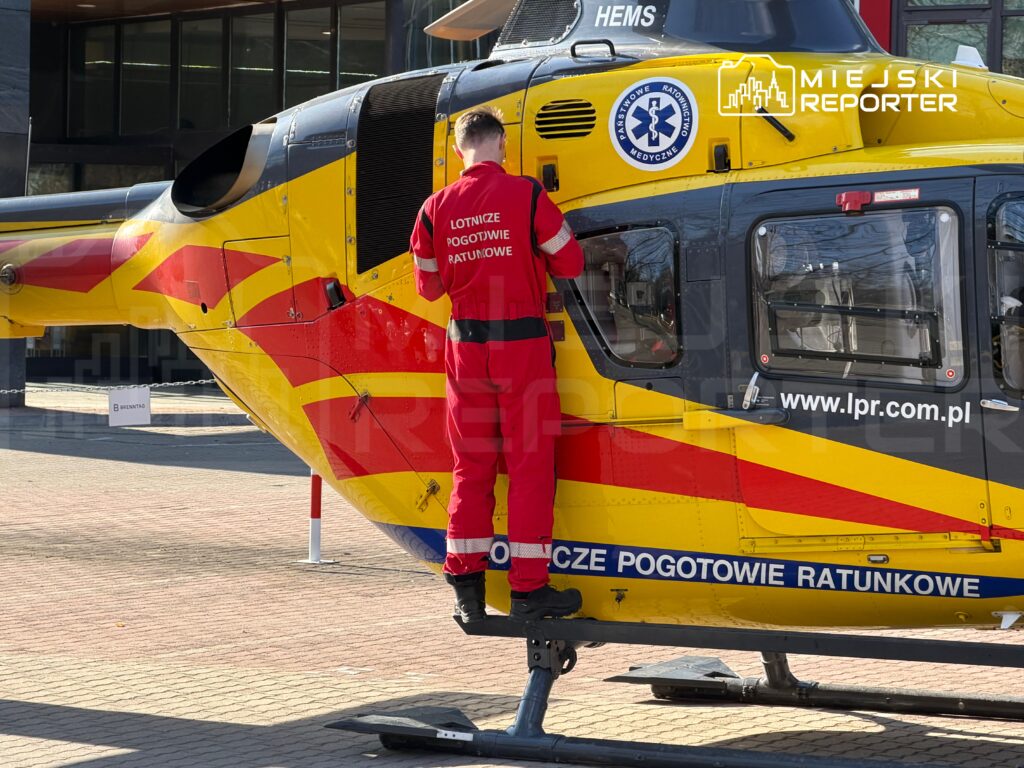 Ratownik medyczny w czerwonym stroju wspina się na pokład helikoptera ratunkowego na placu przed budynkiem.