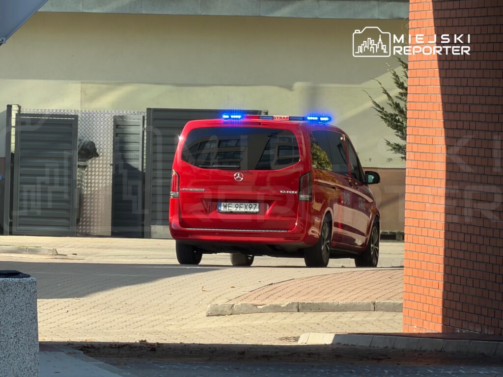 Czerwony samochód służb ratunkowych z niebieskimi światłami, wyjeżdżający z parkingu.