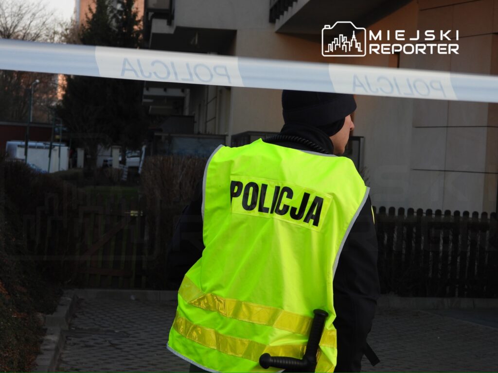 Policjant w żółtej kamizelce ochronnej stoi przy taśmie policyjnej w pobliżu bloku mieszkalnego.