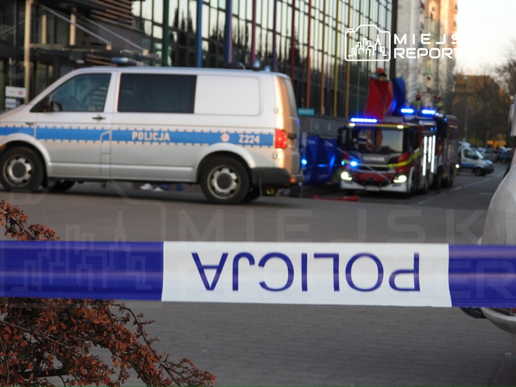 Policyjny radiowóz oraz wóz strażacki zaparkowane w pobliżu miejsca zdarzenia, otoczone taśmą zabezpieczającą.