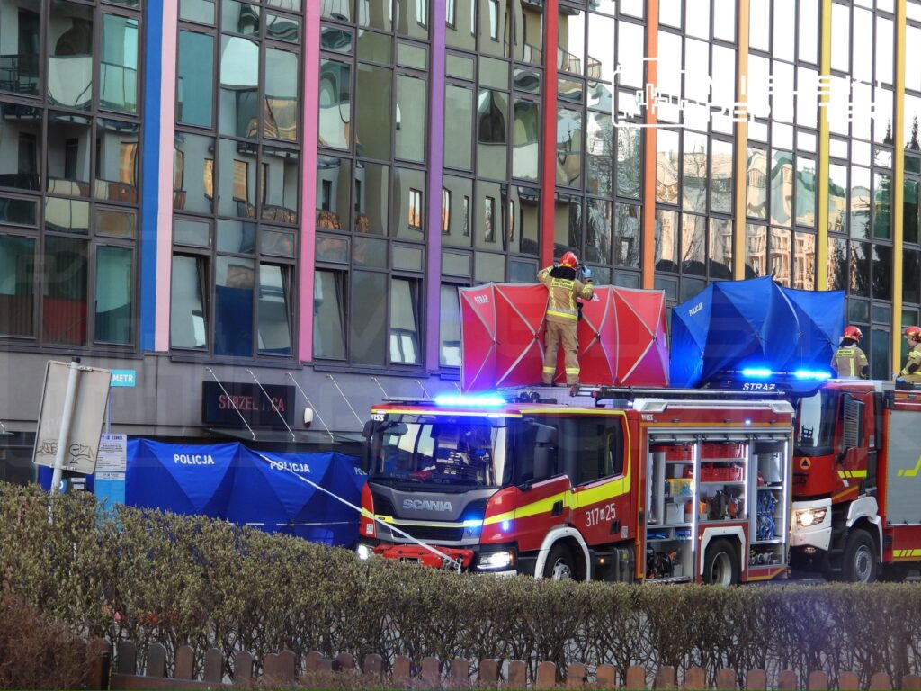 Strażacy pracują przy wozie strażackim, a w tle widać nowoczesny budynek z kolorowymi detalami.