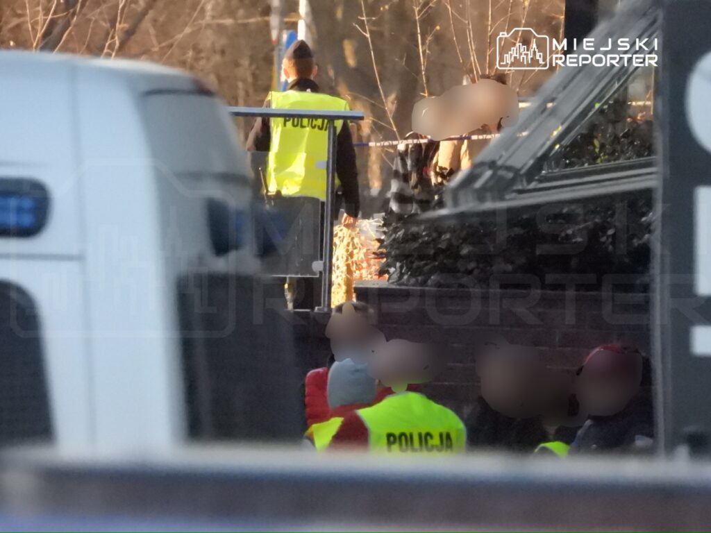 Grupa policjantów w żółtych kamizelkach zabezpiecza teren w pobliżu ogrodzenia, w tle widoczne zarośla.