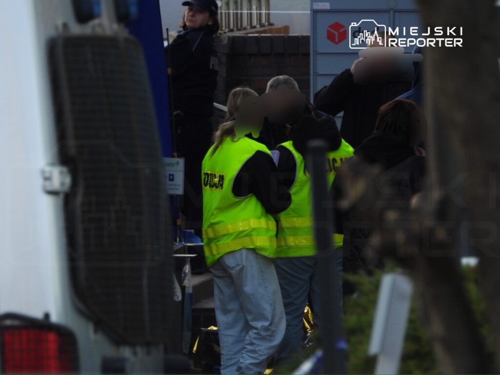 Grupa osób w kamizelkach z napisem "POLICJA" rozmawia obok furgonetki w miejskim otoczeniu.