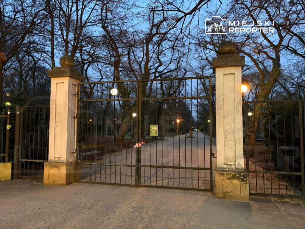 Zamknięte bramy parku, otoczone drzewami, z oświetleniem ulicznym i wyraźnie widoczną alejką.