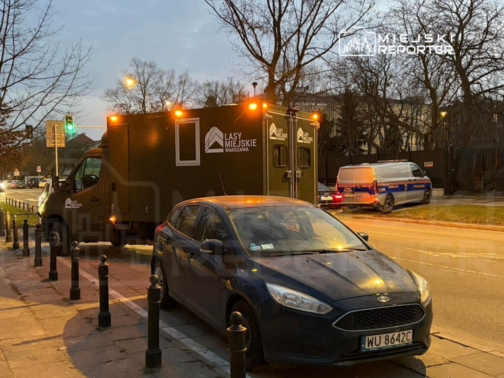 Na ulicy stoi zielony samochód dostawczy z logo "Lasy Miejskie Warszawa", obok zaparkowany niebieski samochód osobowy.