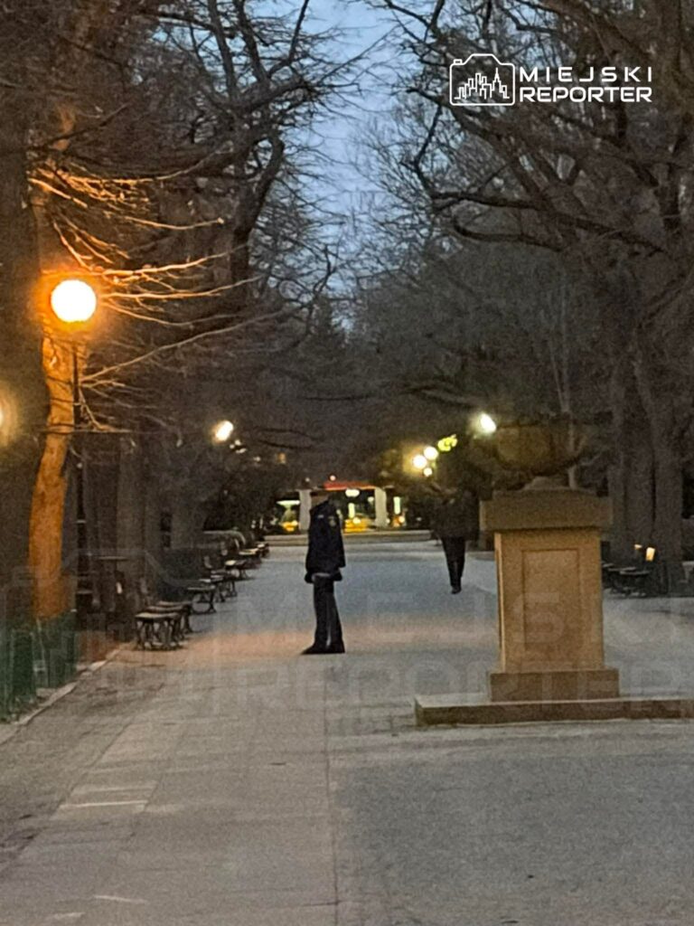 Mężczyzna w mundurze stoi na oświetlonej alei w parku, w tle widać drugą osobę oraz ławki.