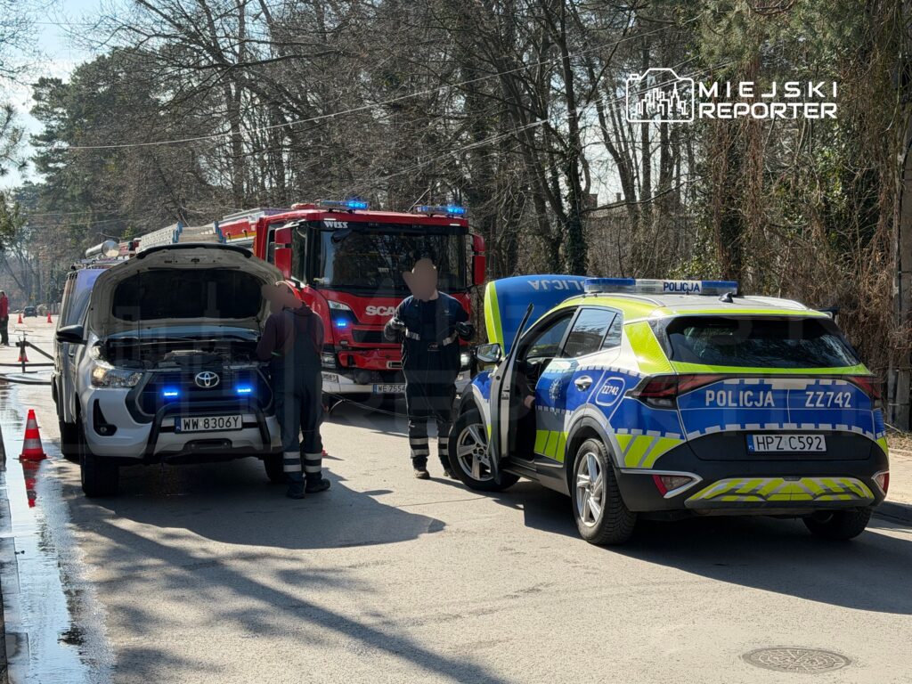 Na ulicy, obok otwartego samochodu terenowego, policjanci sprawdzają jego stan techniczny, a w tle widać wóz strażacki.