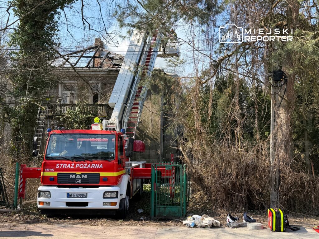 Straż pożarna z drabiną przystosowaną do akcji gaśniczej przy budynku z uszkodzoną dachówką, otoczonym zielenią.