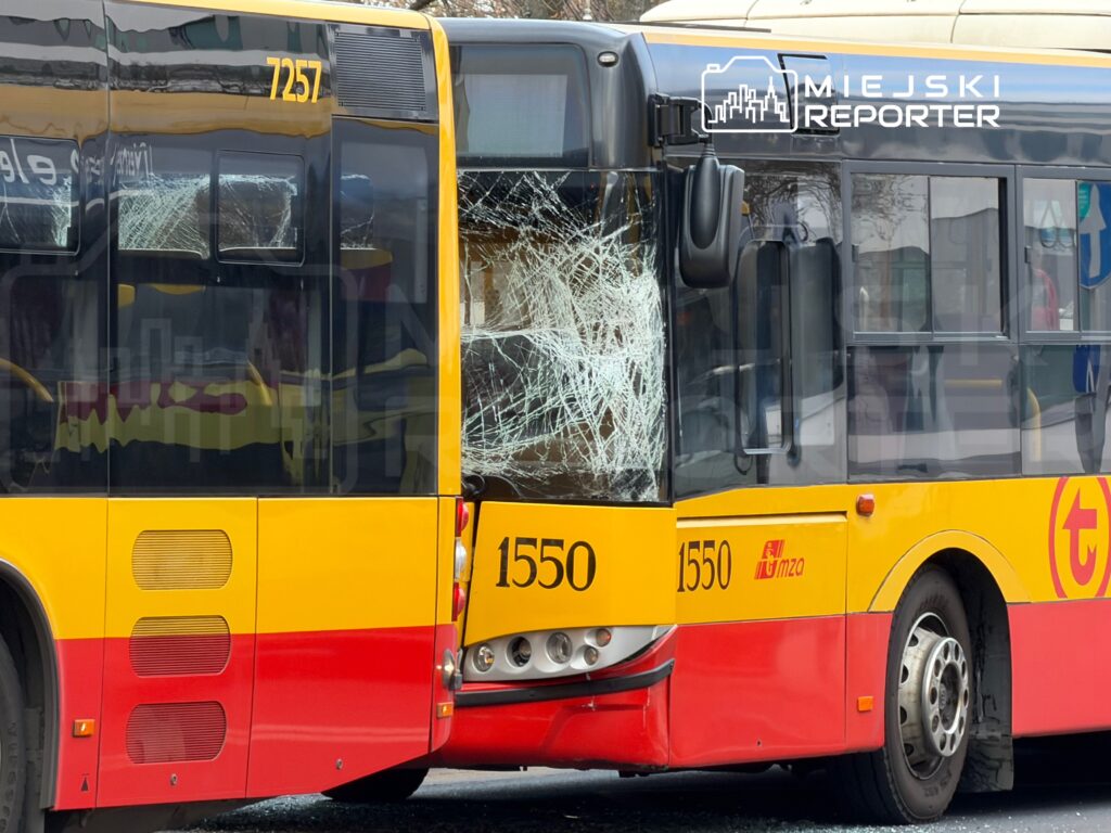 Dwa autobusy miejskie z uszkodzonymi tylnymi szybami stoją obok siebie na przystanku.
