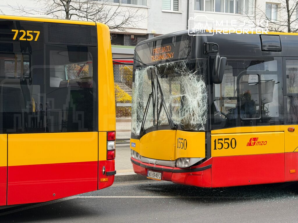 Zniszczony autobus komunikacji miejskiej z pękniętą szybą stoi obok innego autobusu na przystanku.
