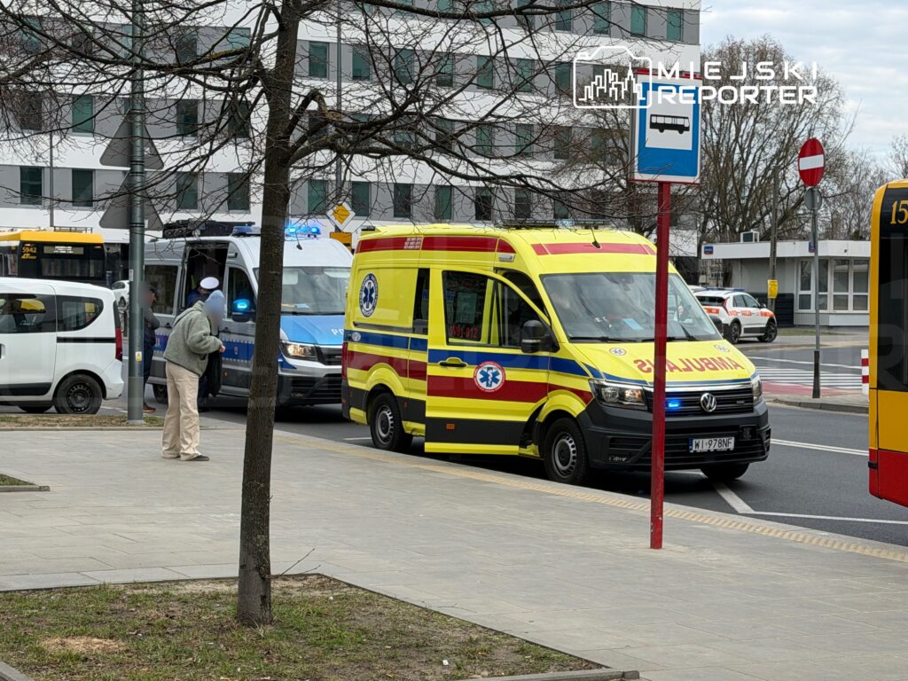 Karetka pogotowia z włączonymi sygnałami świetlnymi zatrzymana na ulicy, obok policjant i przechodzień w szarym swetrze.