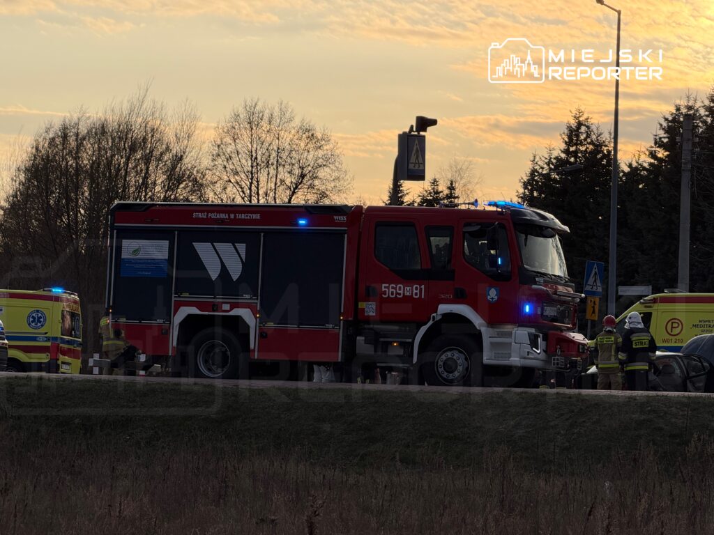 Wozem strażackim z niebieskimi światłami podjeżdża do miejsca zdarzenia, obok stoją ratownicy medyczni.