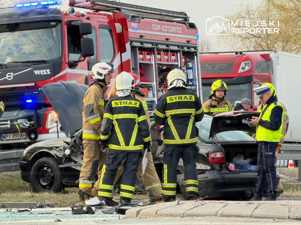 Grupa strażaków w ubraniach ochronnych bada uszkodzony samochód po wypadku drogowym, obok stoi policjant.