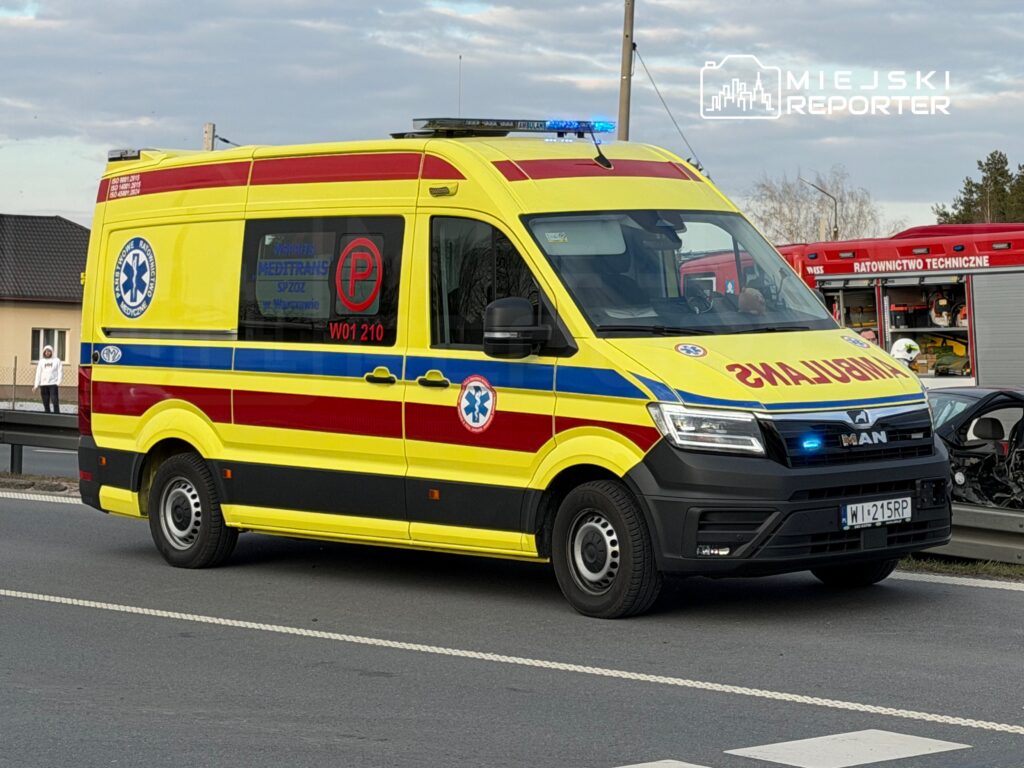 Karetka pogotowia z niebieskimi światłami, zatrzymana na poboczu drogi, obok widać uszkodzony samochód.