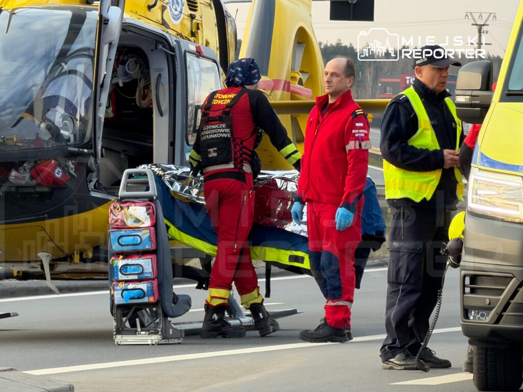 Ratownicy medyczni transportują poszkodowanego na noszach obok śmigłowca lotniczego pogotowia ratunkowego.