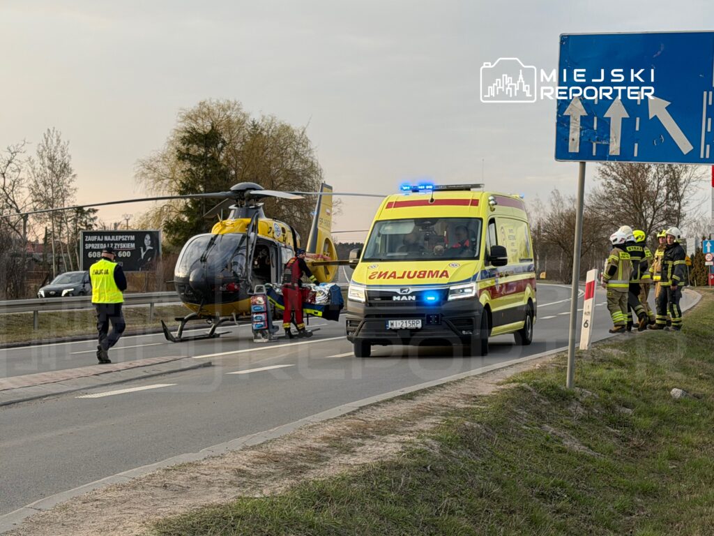 Lądowanie śmigłowca ratunkowego obok karetki, przy drodze, z zespołem ratunkowym w akcji.