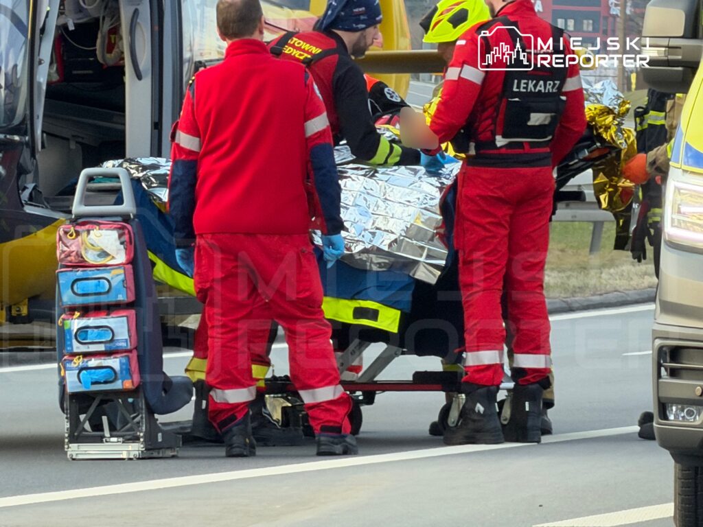 Zespół ratowniczy transportuje poszkodowanego na noszach do karetki na drodze.