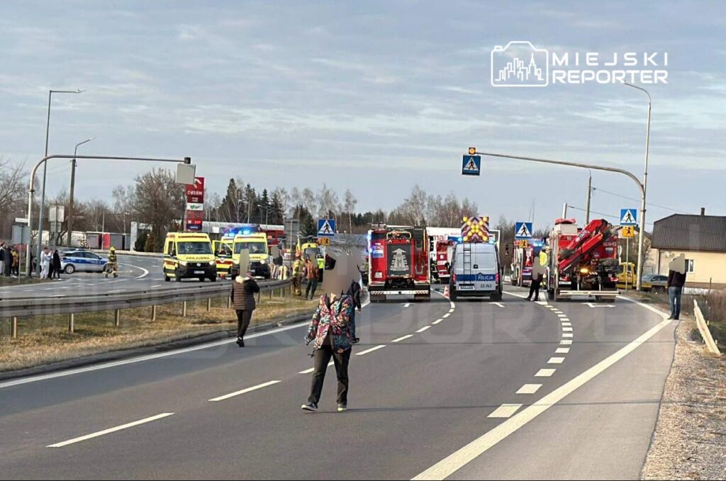 Ratownicy medyczni i strażacy pracują na miejscu wypadku drogowym, obok pojazdów służb ratunkowych.
