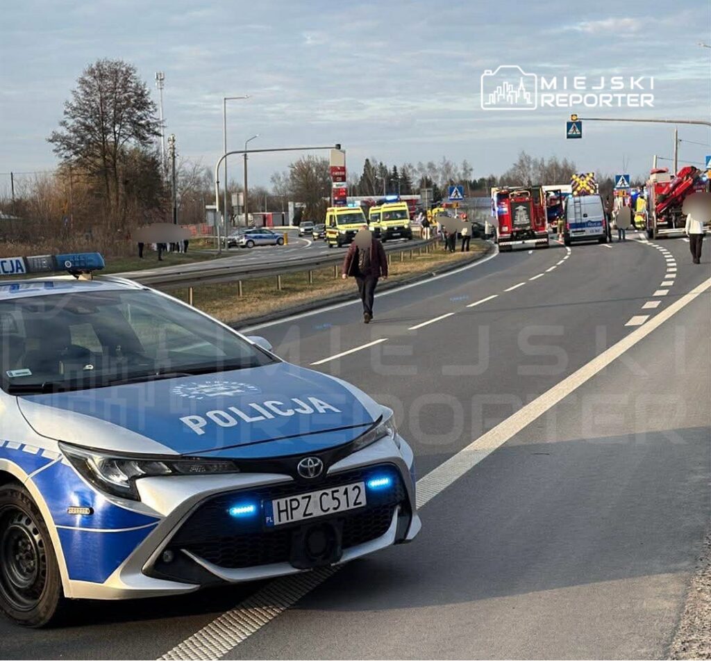 Radiowóz policji stoi na drodze, obok widać kilka pojazdów ratunkowych oraz przechodniów w tle.