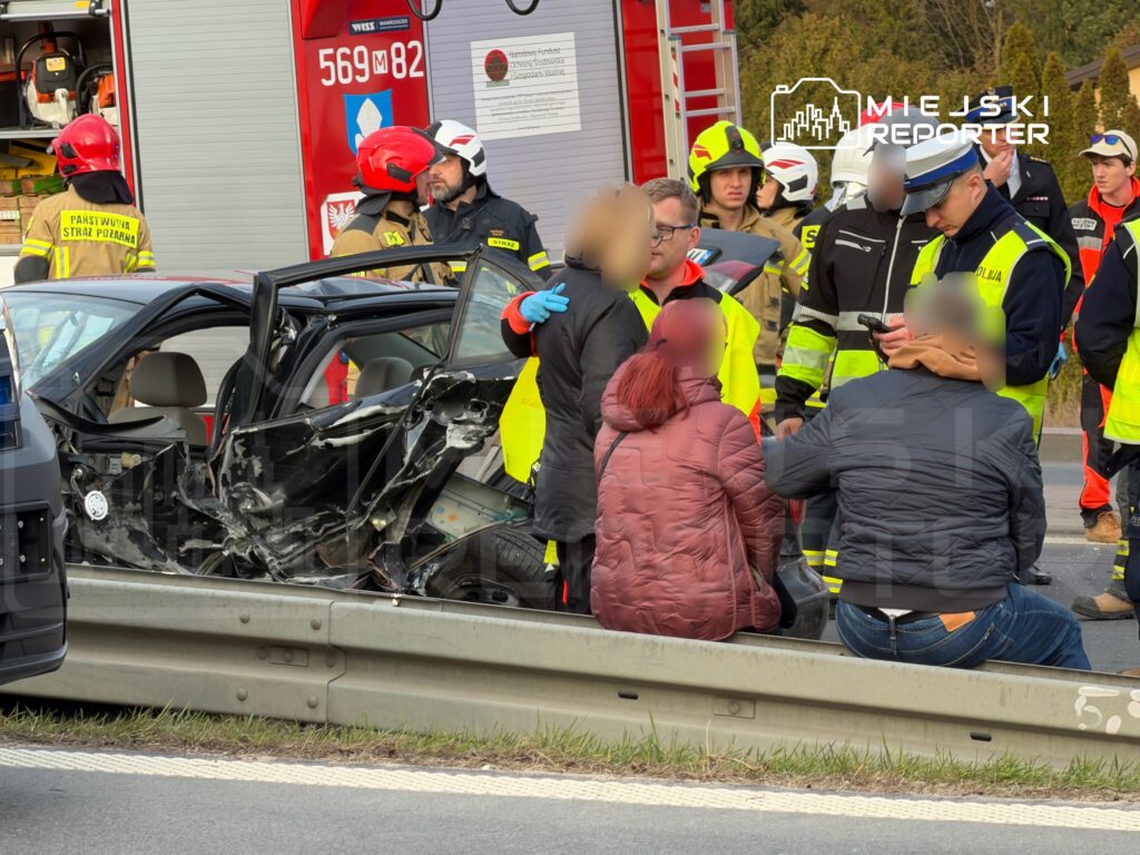 W miejscu wypadku drogowego, ratownicy i policjanci udzielają pomocy poszkodowanym w zniszczonym samochodzie.