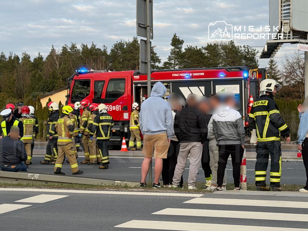 Grupa strażaków w mundurach omawia sytuację obok wozu strażackiego na skrzyżowaniu, a obok zbiera się tłum obserwatorów.