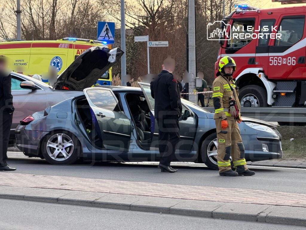 Kierowca w garniturze rozmawia z ratownikami przy uszkodzonym samochodzie, obok stoi wóz strażacki.