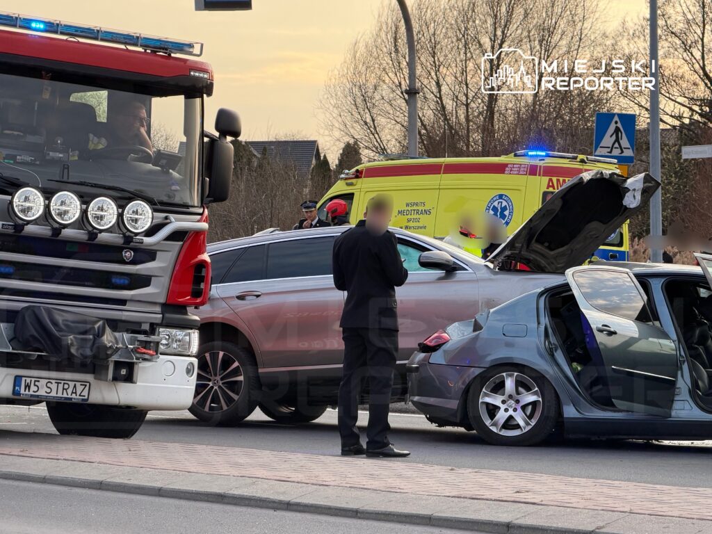W miejscu zdarzenia stoi strażacki wóz z włączonymi sygnałami, obok zniszczony samochód osobowy z otwartymi drzwiami.
