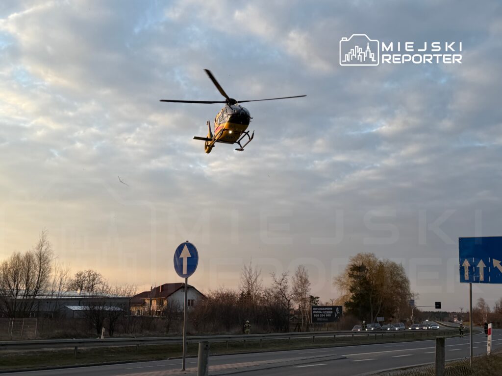Śmigłowiec ratunkowy unosi się nad drogą w pobliżu zabudowań, w tle widoczne chmury i znaki drogowe.