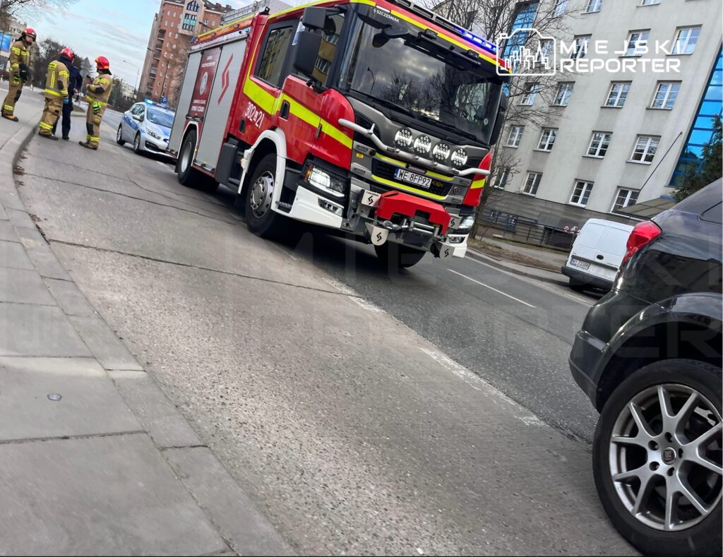 Strażacy rozmawiają obok wozu strażackiego na ruchliwej ulicy, w tle widać zaparkowane samochody.
